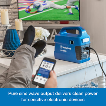 Westinghouse | iGen600s Portable Power Station sits on a table. There are several cords plugged into it. In the foreground someone is holding a phone that is being charged by the power station. A blue banner at the bottom reads,