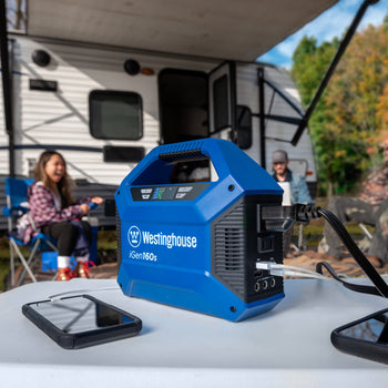 Westinghouse | iGen160s Portable Power Station sitting on a table with a phone charger plugged into it and some other cables plugged in. A group of people are sitting in the background in front of a camper.