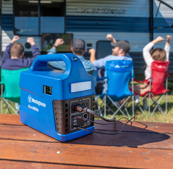 Westinghouse | iGen600s Portable Power Station is shown sitting on a table in the foreground several cables plugged into it. A group of people are watching TV in the background in front of a camper.