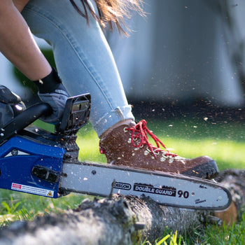 Action shot of a woman cutting a tree branch with a chain saw. The tree branch is laying on the grass and she is holding it down with her boot. Sawdust from the branch is flying in the air.