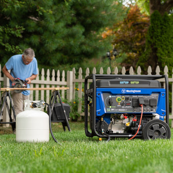 Westinghouse | WGen9500DF portable generator connected to a propane tank sitting in a fenced in yard. A man is sawing wooden boards in the background.
